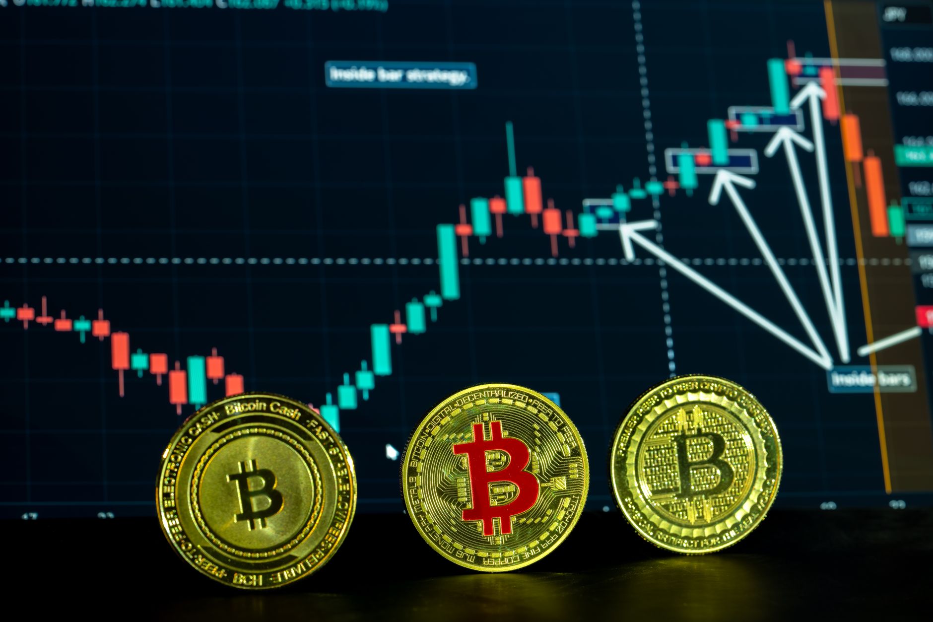 A physical Bitcoin token resting on a desk next to a computer monitor showing a price chart.